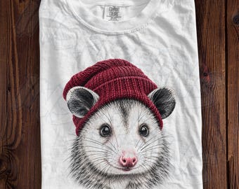 May include: A white crew neck t-shirt featuring a graphic print of an opossum wearing a dark red knit beanie. The shirt has a Comfort Colors brand label printed on the inside of the collar. The design shows the opossum's face with whiskers.