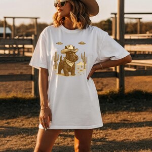 May include: White t-shirt featuring a cartoon illustration of a dog dressed as a cowboy, complete with a hat and bandana. The design includes cacti and UFOs, set against a desert backdrop. A fun and unique graphic tee.