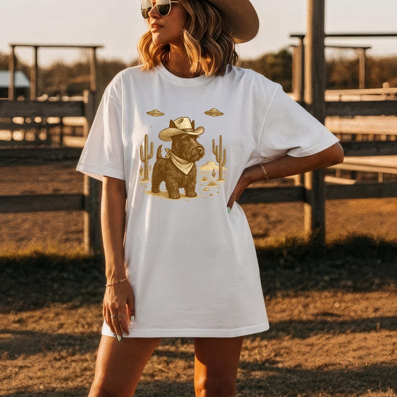May include: White t-shirt featuring a cartoon illustration of a dog dressed as a cowboy, complete with a hat and bandana. The design includes cacti and UFOs, set against a desert backdrop. A fun and unique graphic tee.