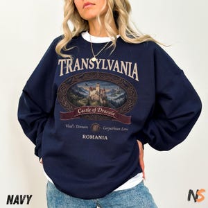 Puede incluir: Una sudadera azul marino de cuello redondo con un estampado gr&aacute;fico de un castillo en un marco ovalado. El texto dice Transylvania, Castle of Dracula, Vlad's Domain, Carpathian Lore y Romania. Se incluye un cuello blanco debajo.