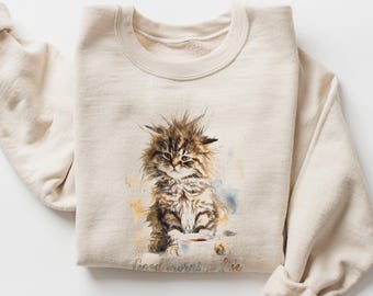 May include: A sand-colored crewneck sweatshirt featuring a watercolor-style illustration of a fluffy, messy-haired kitten sitting next to a small coffee cup. Below the kitten, the text reads Good morning Life in a cursive, brown-toned font.