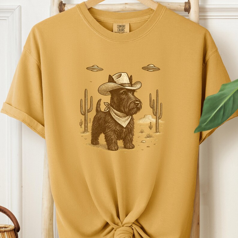 May include: Mustard-colored t-shirt featuring a cartoon illustration of a black dog wearing a cowboy hat and bandana, set against a desert landscape with cacti and UFOs. The shirt has the words "Comfort Colors" printed on the upper right.