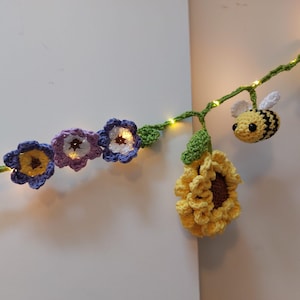 Puede incluir: Guirnalda de ganchillo con flores, un girasol y una abeja. Las flores son de color púrpura, azul y amarillo, con un centro blanco. El girasol es amarillo y marrón, y la abeja es amarilla y negra. La guirnalda tiene pequeñas luces.