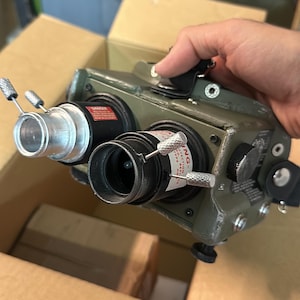 Könnte beinhalten: Ein militärisches Nachtsichtgerät mit einem grünen Metallgehäuse. Das Gerät hat zwei Linsen, mehrere Zifferblätter und silberfarbene Einstellknöpfe. Es wird in einem Karton gehalten.