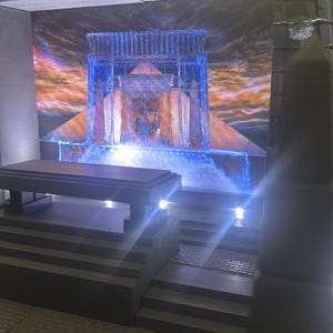Può includere: Un diorama che presenta una struttura in pietra grigio scuro con un altare piatto e gradini. Lo sfondo mostra una scena vibrante e illuminata di un antico tempio con luce blu, nuvole arancioni e obelischi. Include illuminazione a LED integrata.