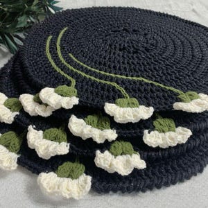May include: A stack of circular black crochet placemats featuring a decorative pattern of green stems and white flower blossoms. Each placemat is crafted with a textured stitch, and the floral elements are arranged along the edges of the mats.