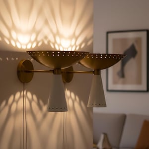 Może przedstawiać: Para kinkietów ściennych w stylu mid-century modern. Każdy kinkiet ma perforowany klosz w kształcie misy w kolorze mosiądzu i białą, stożkową oprawę oświetleniową. Kinkiety są zamontowane na białej ścianie, rzucając wzorzyste światło.