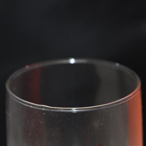May include: A clear, transparent glass with a rounded rim is in the foreground. An orange cylindrical object is in the background. The objects are set against a dark background. The glass appears to be empty.