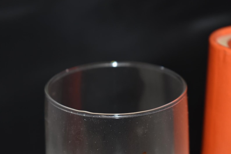May include: A clear, transparent glass with a rounded rim is in the foreground. An orange cylindrical object is in the background. The objects are set against a dark background. The glass appears to be empty.