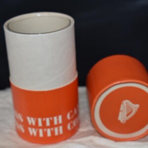 May include: Two cylindrical containers, one upright and one on its side. The containers are orange with white tops. Text on the side of the upright container reads "WITH CA... WITH CO...". The bottom of the container on its side has a white circle with a harp symbol.