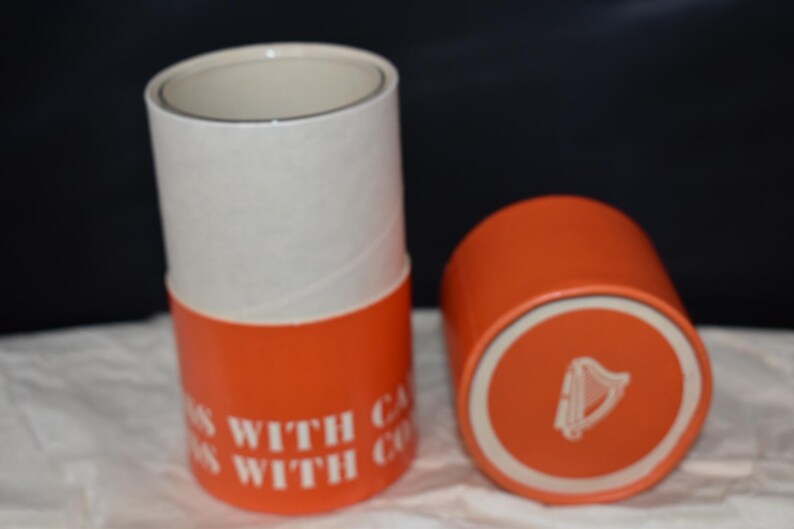 May include: Two cylindrical containers, one upright and one on its side. The containers are orange with white tops. Text on the side of the upright container reads "WITH CA... WITH CO...". The bottom of the container on its side has a white circle with a harp symbol.