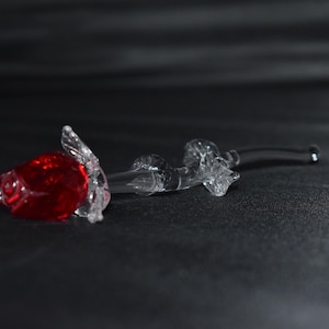 May include: A glass rose sculpture featuring a bright red bloom and clear stem and leaves. The delicate glasswork captures the form of a rose, creating a decorative piece. The rose is lying on a dark surface.