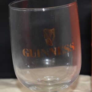May include: A clear stemmed glass with the word "GUINNESS" in gold lettering and a gold harp logo. The glass is partially in view, with an orange cylindrical container visible in the background.