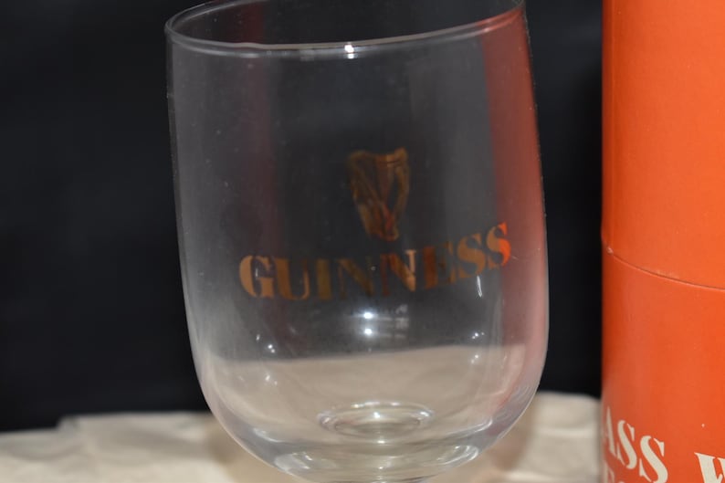 May include: A clear stemmed glass with the word "GUINNESS" in gold lettering and a gold harp logo. The glass is partially in view, with an orange cylindrical container visible in the background.