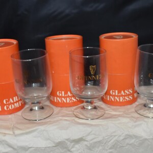 May include: Clear stemmed glasses with the Guinness logo in gold, displayed with orange cylindrical boxes. The boxes have white text that reads "GLASS GUINNESS". The glasses have a clear bowl and a silver base.