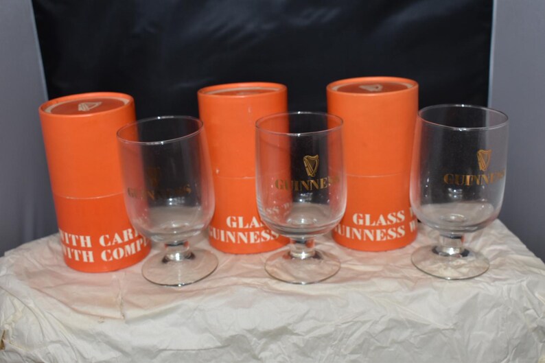 May include: Clear stemmed glasses with the Guinness logo in gold, displayed with orange cylindrical boxes. The boxes have white text that reads "GLASS GUINNESS". The glasses have a clear bowl and a silver base.