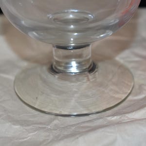 May include: A clear glass goblet with a rounded bowl and a circular base. The stem is short and connects the bowl to the base. The goblet is sitting on a white, textured surface. An orange container with white text is visible in the background.