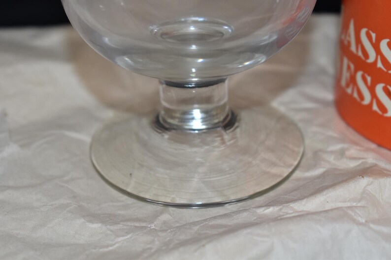 May include: A clear glass goblet with a rounded bowl and a circular base. The stem is short and connects the bowl to the base. The goblet is sitting on a white, textured surface. An orange container with white text is visible in the background.