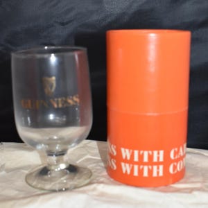 May include: A clear Guinness glass with a gold logo and a stem, next to an orange cylinder with white text. The glass has a rounded bowl shape. The cylinder has the text "S WITH CA" and "S WITH CO".