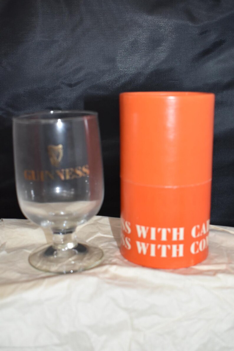May include: A clear Guinness glass with a gold logo and a stem, next to an orange cylinder with white text. The glass has a rounded bowl shape. The cylinder has the text "S WITH CA" and "S WITH CO".