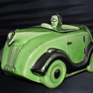 May include: A green ceramic vintage car figurine with black accents. The car has a driver figure wearing glasses and a helmet. The wheels and fenders are black. The license plate reads OK 757.