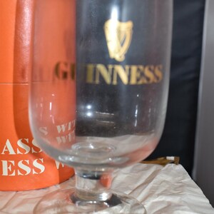 May include: A clear glass Guinness pint glass with a wide base and a curved body. The Guinness logo is in gold on a black background. An orange box is partially visible behind the glass, with white text.