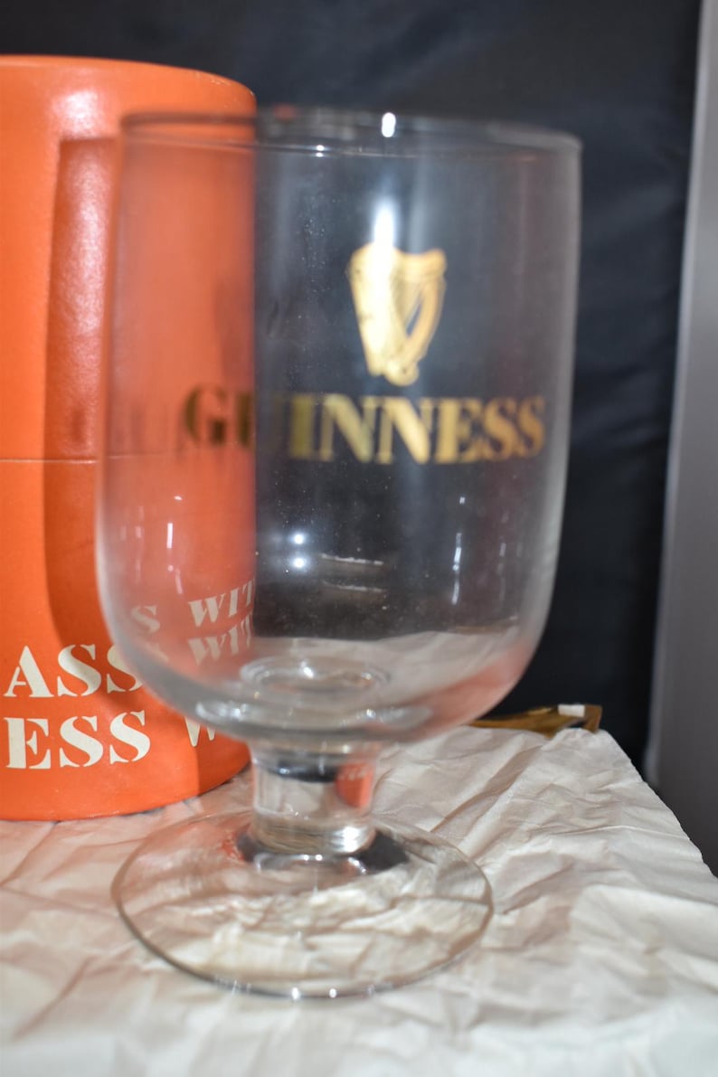 May include: A clear glass Guinness pint glass with a wide base and a curved body. The Guinness logo is in gold on a black background. An orange box is partially visible behind the glass, with white text.