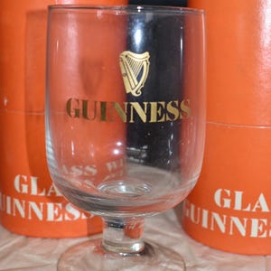 May include: A clear Guinness glass with a gold harp logo and the word "GUINNESS" in gold lettering. The glass is in front of two orange cylindrical boxes with the words "GUINNESS" printed on them.