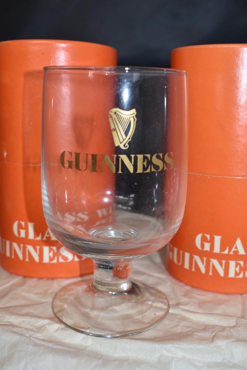 May include: A clear Guinness glass with a gold harp logo and the word "GUINNESS" in gold lettering. The glass is in front of two orange cylindrical boxes with the words "GUINNESS" printed on them.