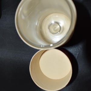 May include: An overhead shot of a clear glass with a rounded rim and a cylindrical container. The glass is empty, and the container is open, revealing a beige interior. The background is a dark, solid color.