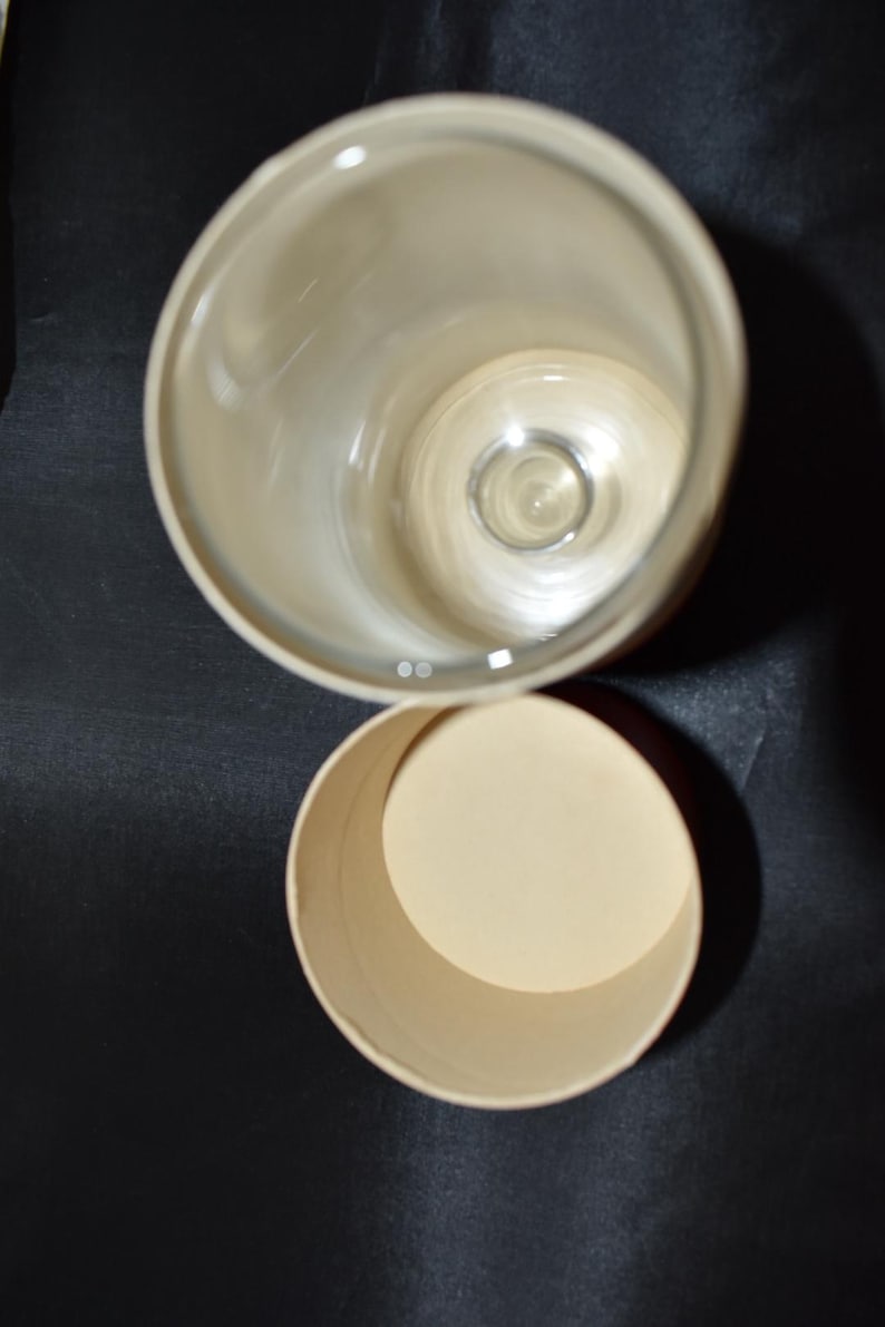 May include: An overhead shot of a clear glass with a rounded rim and a cylindrical container. The glass is empty, and the container is open, revealing a beige interior. The background is a dark, solid color.