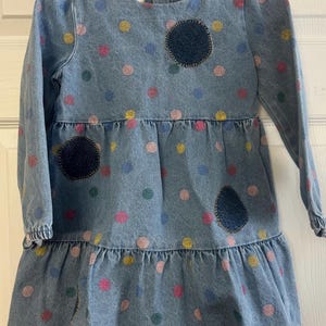 May include: A light blue denim dress with long sleeves and a ruffled hem. The dress is decorated with colorful polka dots and three dark blue patches. The dress is hanging on a white hanger.
