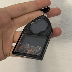 Peut inclure: Un porte-clés shaker en acrylique noir en forme de fenêtre gothique en arc. Le cadre présente des motifs célestes gravés, notamment des étoiles et des lunes. À l'intérieur du compartiment transparent, des breloques illustrées bougent.