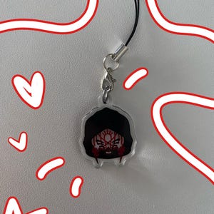 May include: A clear acrylic charm featuring a stylised character head with black hair and a red and white patterned mask. The charm is attached to a silver-toned metal lobster clasp and a thin black cord loop, accented by red hand-drawn digital doodles.