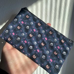 May include: A navy blue zippered pouch with a repeating pattern of skulls, pink hearts, and pink rectangles. The skulls are white and the faces are peach-colored. The pouch is held in a hand.