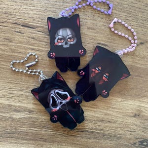 May include: Three black cat-shaped keychains with spooky designs. One features a skull, another a Scream mask, and the third has red eyes. Each keychain has a colored beaded chain for attachment.