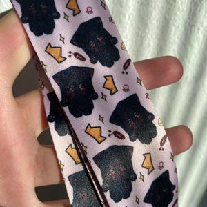 May include: A light pink lanyard with a repeating pattern of black bear-like figures, gold crowns, and small pink paw prints. The lanyard is made of a smooth, possibly satin, material.