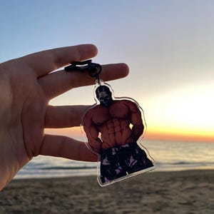 May include: A clear acrylic keychain featuring a muscular figure with a black mask and patterned shorts. The keychain is held up against a beach and sunset backdrop, with the ocean visible in the distance.
