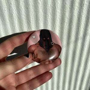 Puede incluir: Un botón redondo rosa con una ilustración de dibujos animados de una persona con un pasamontañas negro. La ilustración incluye un corazón y una pequeña cadena dorada. El botón se sostiene en una mano.