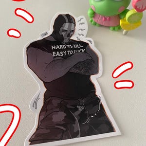 Puede incluir: Una pegatina con una ilustración estilizada de un personaje con máscara negra y equipo táctico, con el texto "HARD TO KILL EASY TO F*CK" en la camiseta. Una figura de rana verde está en el fondo.