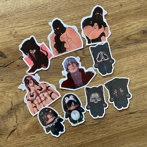 Puede incluir: Una colección de pegatinas variadas con ilustraciones de estilo anime. Las pegatinas representan personajes en diferentes poses y disfraces, incluyendo un pingüino, un gato y figuras con máscaras. Las pegatinas están sobre una superficie de madera.