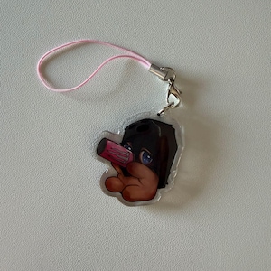 Op de afbeelding: Een acryl bedel met een cartoonfiguur met donker haar en blauwe ogen, die een zwart masker draagt en een roze drankblikje vasthoudt. De bedel is bevestigd aan een zilverkleurige metalen karabijnhaak en een dun, lichtroze nylon luskoord.