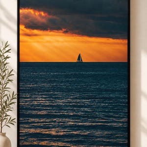 Op de afbeelding: Een ingelijste muurkunstprint met een zeilboot op een donkerblauwe oceaan onder een levendige oranje zonsonderganghemel met dramatische wolken. De horizonlijn scheidt het diepe water van de gloeiende atmosfeer in dit landschap.