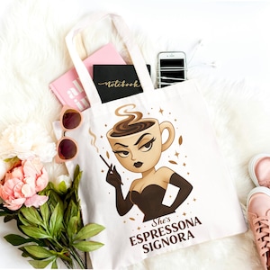 May include: A white tote bag with a coffee-themed illustration. The design features a woman's face inside a coffee cup, holding a cigarette. The text "She's Espressona Signora" is below the image. The bag is filled with notebooks and a phone.