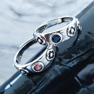 May include: Two silver rings with floral designs and gemstone accents. One ring features a deep blue stone, the other a red stone. The rings are set against a dark, reflective surface.