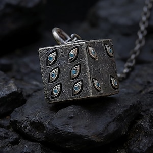 May include: A silver-toned pendant necklace featuring a cube-shaped charm. The charm is adorned with eye-shaped designs, each containing a small, light blue stone. The necklace chain is also silver-toned, and the pendant rests on a dark, textured surface.