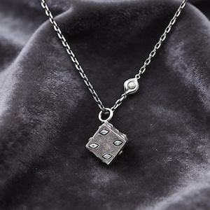 May include: A silver necklace with a dice pendant. The dice is square with four blue-green gemstones. The chain is delicate and hangs on a dark gray velvet background. The necklace has a small, round accent near the clasp.