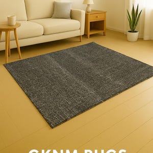 May include: A rectangular, dark gray rug with a textured, striped pattern. The rug is placed on a light wood floor in a living room setting. The text "GKNM RUGS" is displayed at the bottom of the image.