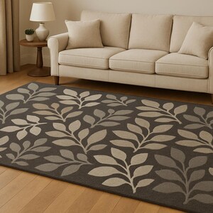 Puede incluir: Una alfombra rectangular con un diseño de hojas en tonos grises y beige. La alfombra está colocada sobre un suelo de madera frente a un sofá beige. Las hojas están dispuestas en un patrón repetitivo.