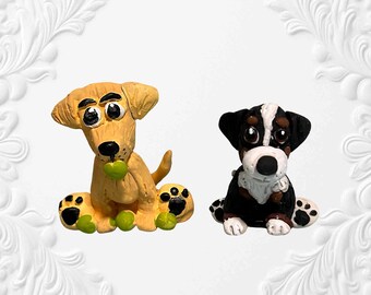 May include: Two handmade dog figurines. One is a light yellow Labrador with a green ball in its mouth. The other is a black, white, and brown dog. Both have large eyes and paw prints on their feet.